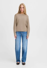 Pull en maille beige avec un col rond et des manches longues, associé à un jean bleu clair à jambes larges et des chaussures plates noires.