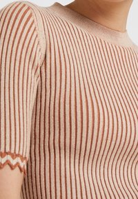 Gebreide top met horizontale strepen in lichtbeige en roest. Heeft een ronde halslijn en golvende randen bij de mouwen. Zachte textuur.