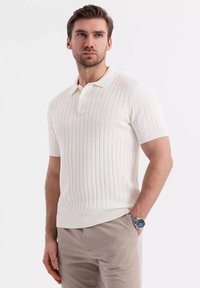 Bílá pruhovaná polo košile s krátkým rukávem, klasickým límečkem a zapínáním na tři knoflíky, v kombinaci s světle béžovými kalhotami a stříbrnými hodinkami.