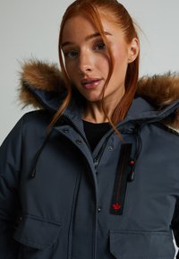 Jūras spēku pūkotā jakas ar kažokādas apdari, divām priekšējām kabatām un vertikālu melnu paneli ar sarkaniem metāla elementiem un lapas emblēmu.