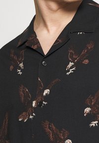 Schwarze Hemdbluse mit braunen und weißen Adlerdrucks, getragen von einer Person, die den Hals und den oberen Brustbereich zeigt.