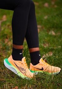 Personne portant des chaussures de course Nike orange vif et vert avec des chaussettes noires Nike, se tenant sur de l'herbe.