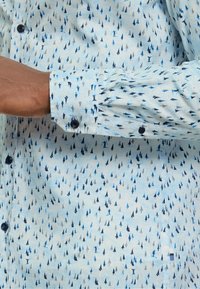 Helles blaues Langarmhemd mit einem Muster aus kleinen schwarzen, grauen und blauen Bäumen. Knöpfe an den Manschetten mit einer glatten Textur.