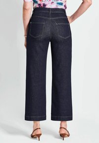 Donkerblauwe denim wijde broek met twee achterzakken en contrasterende gouden stiksel; model draagt bruine hakken.