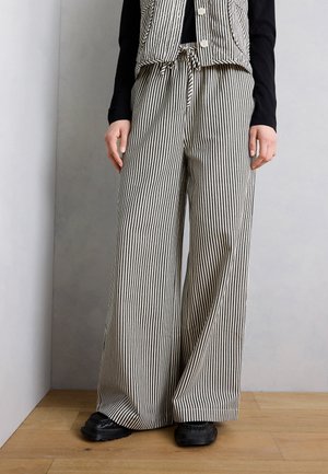 Άτομο που φοράει παντελόνι με πλατύ πόδι και κάθετες ασπρόμαυρες ρίγες, με αντίστοιχο γιλέκο, μαύρο μπλουζάκι μακρύ μανίκι και μαύρα παπούτσια, σε ξύλινο πάτωμα.