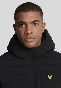 Zwarte gewatteerde capuchonjas met ritssluiting aan de voorkant en een klein geel logo links op de borst. Soepele stof met een lichtgewicht ontwerp.