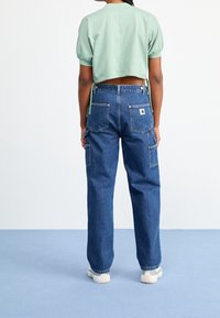 Persona vistiendo jeans de mezclilla azules de tipo carpintero con bolsillos de parche y una camiseta corta de manga corta de color verde claro, de pie sobre un suelo azul claro.