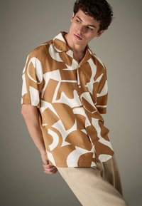 Trumpomis rankovėmis marškiniai rudos spalvos su baltais abstrakčiais raštais; apykaklės dizainas, lengvas audinys, laisvas siluetas, užsegami mygtukais.