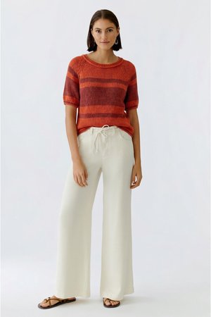 Vrouw draagt een kort mouw oranje en rood gestreepte gebreide top, witte broek met wijde pijpen en trekkoord, en bruine platte sandalen, staand op een witte achtergrond.