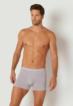 Graue Baumwoll-Boxershorts mit einem weichen Bund und einer taillierten Passform, getragen von einem Mann ohne Hemd. Glatte Textur und kurze Länge.