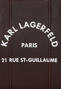 Ruda tekstūruota oda su įspaustu baltu tekstu: "KARL LAGERFELD," "PARIS," ir "21 RUE ST-GUILLAUME." Paprastas, modernus dizainas.