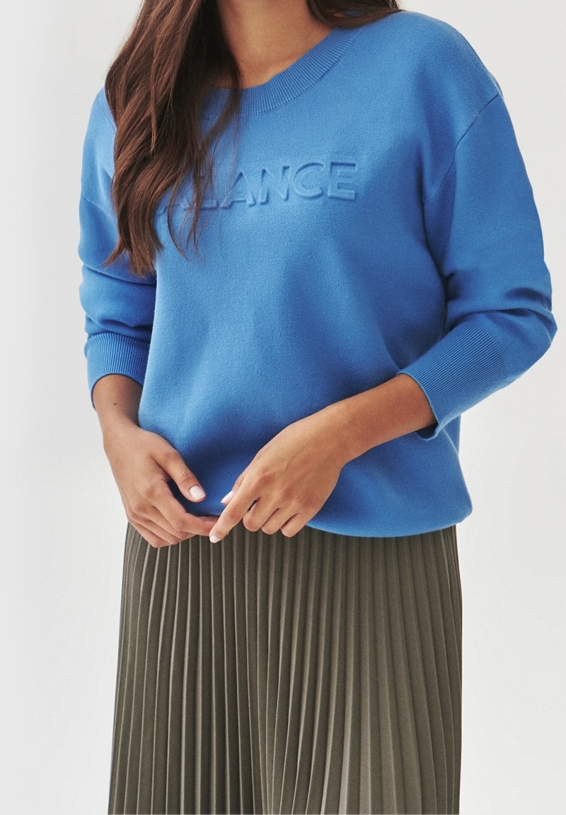 Femme portant un pull bleu avec le texte embossé « BALANCE » et une jupe plissée vert olive, les mains délicatement jointes au niveau de la taille.