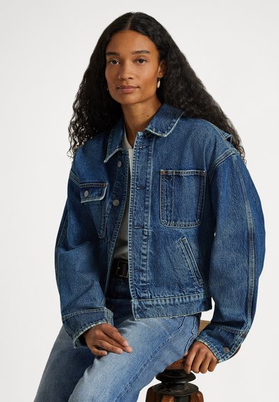 Kvinne med langt, krøllete hår som har på seg en blå denimjakke og jeans, sittende på en trekrakk mot en ensfarget bakgrunn.
