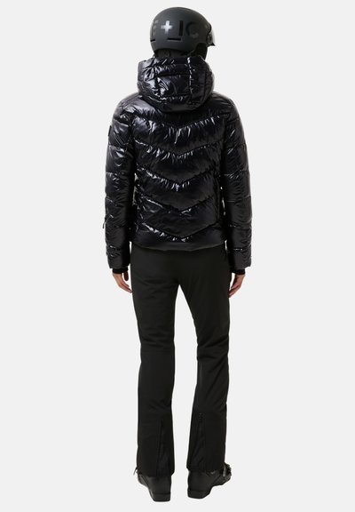 Zwarte pufferjas met capuchon met chevron-stiksels, glanzende afwerking en een getailleerd ontwerp, gecombineerd met zwarte skibroeken en laarzen.