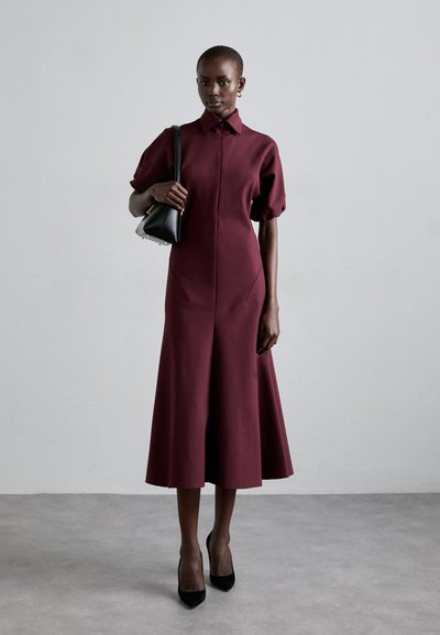 Rochie midi burgundy cu guler tip cămașă, mâneci bufante și buzunare laterale. Purtată cu tocuri negre și o geantă mică neagră.