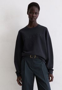 Marineblauwe sweatshirt met reliëf "Marc O'Polo" logo, ronde halslijn en oversized pasvorm, gecombineerd met op maat gemaakte donkergroene broeken en een zwarte riem.