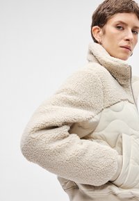 Beige bolyhos dzseki textúrált külsővel, magas gallérral és steppelt részekkel. Nyitott elülső résszel és bordázott mandzsettákkal rendelkezik.