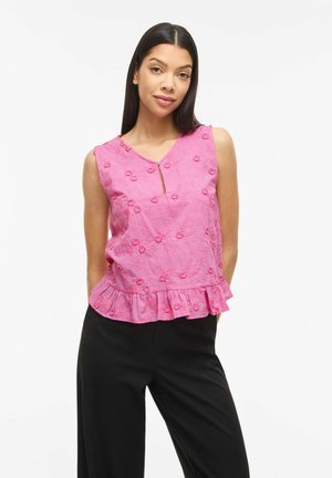 Kobieta o długich ciemnych włosach nosi różowy haftowany top bez rękawów z falbankowanym dołem i czarne spodnie, stoi na białym tle.