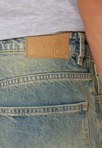 Jeans en denim bleu clair avec une patch en cuir étiqueté "YOURTURN" à l'arrière, avec des coutures visibles et des détails de poches.