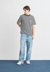 Ριγέ μαύρο και λευκό t-shirt με στρογγυλή λαιμόκοψη και μικρό λογότυπο στο στήθος, σε συνδυασμό με ανοιχτό μπλε φαρδιά τζιν και λευκά αθλητικά παπούτσια.