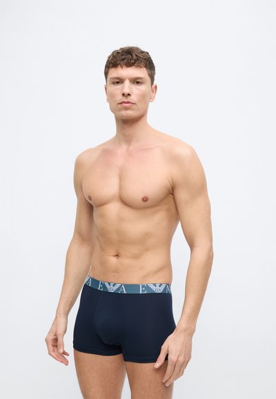 Άνδρας χωρίς μπλούζα με κοντά, σγουρά μαλλιά, φοράει μπλε ναυτικό μπόξερ με μπλε ελαστική ζώνη που έχει λευκό λογότυπο EA, στέκεται μπροστά σε απλό φόντο.