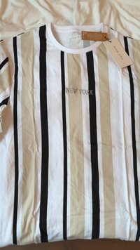 T-shirt s črtami, ki prikazujejo navpične črne, bele in bež črte, z vezenim napisom "NEW YORK" na sprednji strani. Mehka tkanina z oznako.