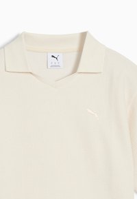 Ljusbeige polo med en strukturerad väv, V-ringning och en liten broderad logotyp på vänster bröst. Klassisk kragsstil.