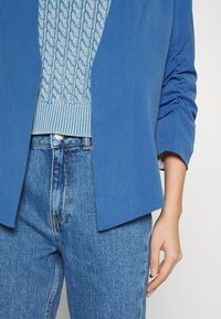 Modrý na míru ušitý blazer má hladkou texturu, s nařasenými rukávy a otevřeným předním dílem. Nošený přes světle modrý svetr s copánkovým vzorem a džínové kalhoty.