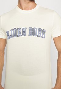 Krämfärgad kortärmad T-shirt med "BJÖRN BORG" i stora blå bokstäver över bröstet, tillverkad av mjukt tyg med en slät yta.