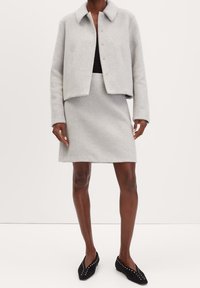 Femme portant une veste en laine gris clair et une jupe assortie avec un haut noir et des ballerines noires cloutées sur un fond uni.
