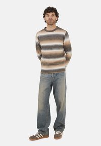 Rustykalny sweter w paski z miękkiego materiału, w odcieniach brązu, beżu i bieli; w połączeniu z jasnoniebieskimi dżinsami i ciemnymi trampkami.