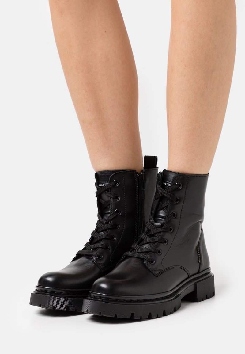 Bottes basses en cuir noir à lacets avec semelles épaisses en caoutchouc portées sur des jambes nues sur un fond blanc.