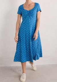 Blå klänning med korta ärmar, samlad mitt, vita prickar och en flödande kjol. Bärs med ljusa sneakers. Slät tygtextur.