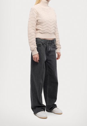 Person iført en cremefarvet strikket rullekravesweater, løstsiddende mørkegrå jeans og hvide sneakers, stående mod en ensartet lys baggrund.