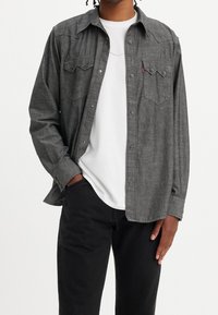 Camicia in flanella grigia con chiusura frontale a bottoni, due tasche sul petto e maniche lunghe. Indossata sopra una t-shirt bianca con scollo rotondo.