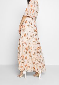 Robe maxi florale en beige clair avec des motifs orange et verts. Elle présente des manches courtes, une silhouette fluide et un ourlet à volants. Associée à des escarpins blancs.