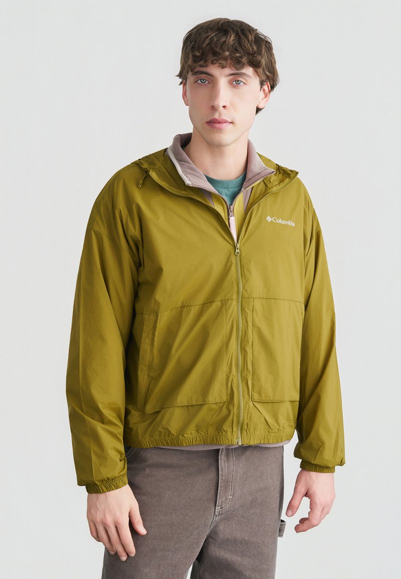 Νεαρός άνδρας με κοντά σγουρά μαλλιά, φοράει μια ζακέτα Columbia σε χρώμα ελιάς με φερμουάρ και γκρι παντελόνι, στέκεται μπροστά σε απλό λευκό φόντο.