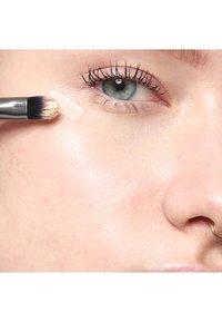 Gros plan d'une personne appliquant un correcteur beige sous l'œil avec un pinceau de maquillage, près d'un anneau de nez, sur une peau claire.