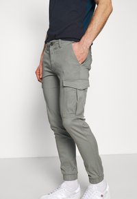 Homme portant un pantalon cargo gris slim avec des poignets élastiques, des poches latérales à rabat, une ceinture noire, des chaussettes blanches et des baskets blanches, se tenant de profil.