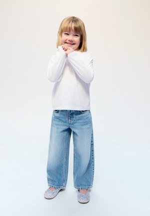 Nasmejana mlada deklica s svetlimi lasmi, oblečena v belo majico z dolgimi rokavi, široke modre kavbojke in bleščeče srebrne čevlje, stoji pred enobarvnim ozadjem.