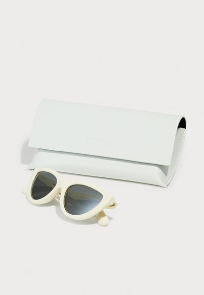 Sunglasses ve tvaru kočičích očí s bledě žlutými rámy a tmavými skly vedle bílého štíhlého hranatého pouzdra označeného značkou "Burberry".