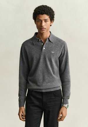 Jeune homme aux cheveux courts et bouclés, portant un pull polo gris à manches longues et un pantalon noir, debout devant un fond clair uni.