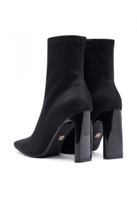 Botas de tobillo negras hechas de tela suave, con punta afilada, un tacón grueso de charol y detalles de costura visibles en los lados.
