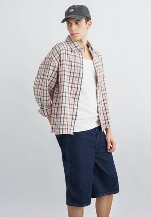 Junger Mann mit schwarzer Kappe, rosa-grau kariertem Hemd über einem weißen Tanktop und dunkelblauen, knielangen Jeansshorts, seitlich stehend.