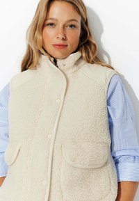 Gilet en polaire beige avec un col haut, fermeture à pression à l'avant et deux poches avant. Présente un tissu texturé et une apparence douce et confortable.
