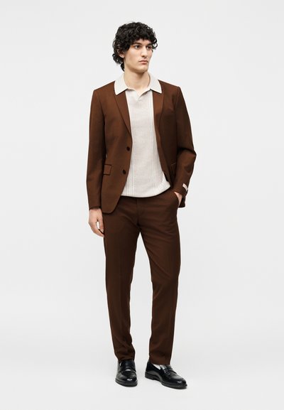 Άνδρας με σγουρά σκούρα μαλλιά φοράει καφέ κοστούμι με λευκό πλεκτό πόλο και μαύρα δερμάτινα μοκασίνια, στέκεται με το ένα χέρι στην τσέπη.