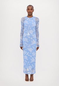 Robe longue à manches longues bleue avec imprimé floral et accents à franges, fabriquée à partir d'un tissu léger et transparent, tombant jusqu'aux chevilles. Talons ouverts marron.
