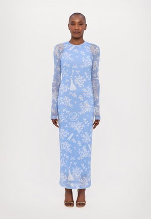 Blue long-sleeve dress with floral print and tassel accents, made from a lightweight sheer fabric, flowing to the ankles. Brown open-toe heels.