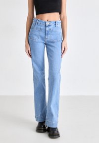 Ljust blå högmidjade jeans med rak bendesign, knappstängning och två bakfickor, i kombination med svarta ankelskorskor.