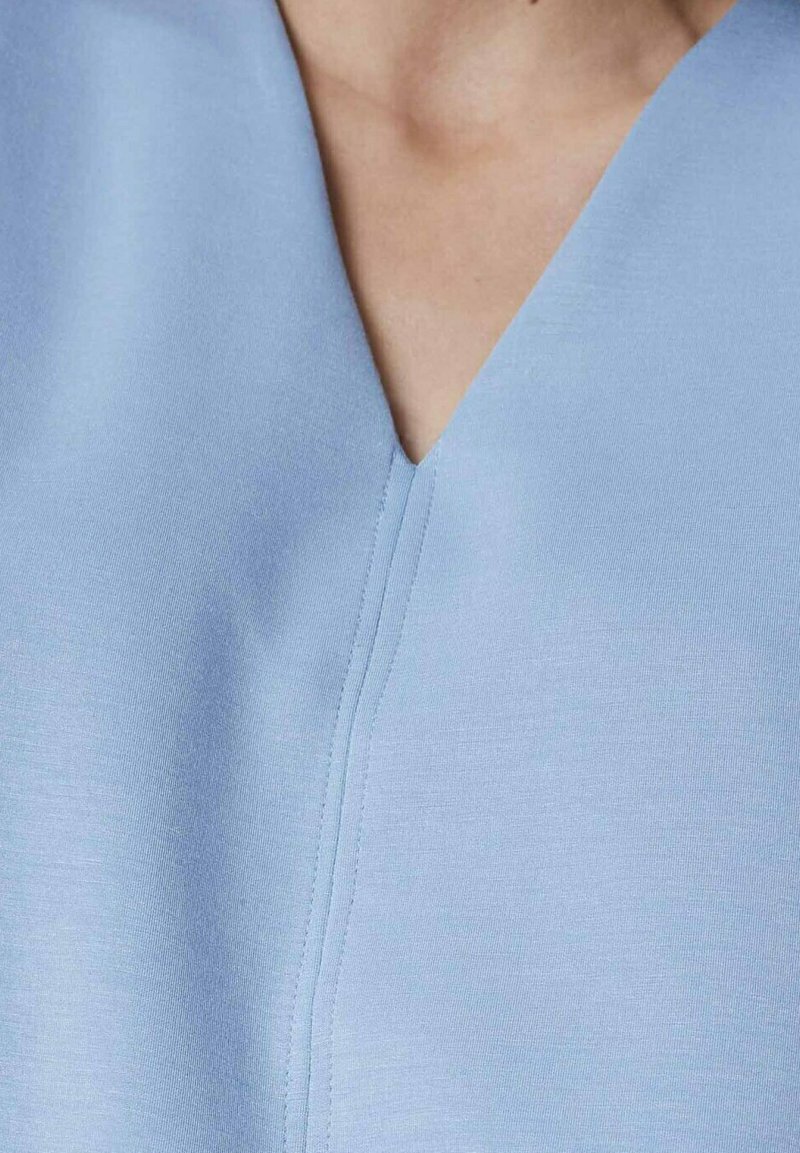 Himmelblåt stof med en glat tekstur, der har en V-hals-design og synlige sømme langs knaplisten. Nærbillede af beklædningsgenstanden.